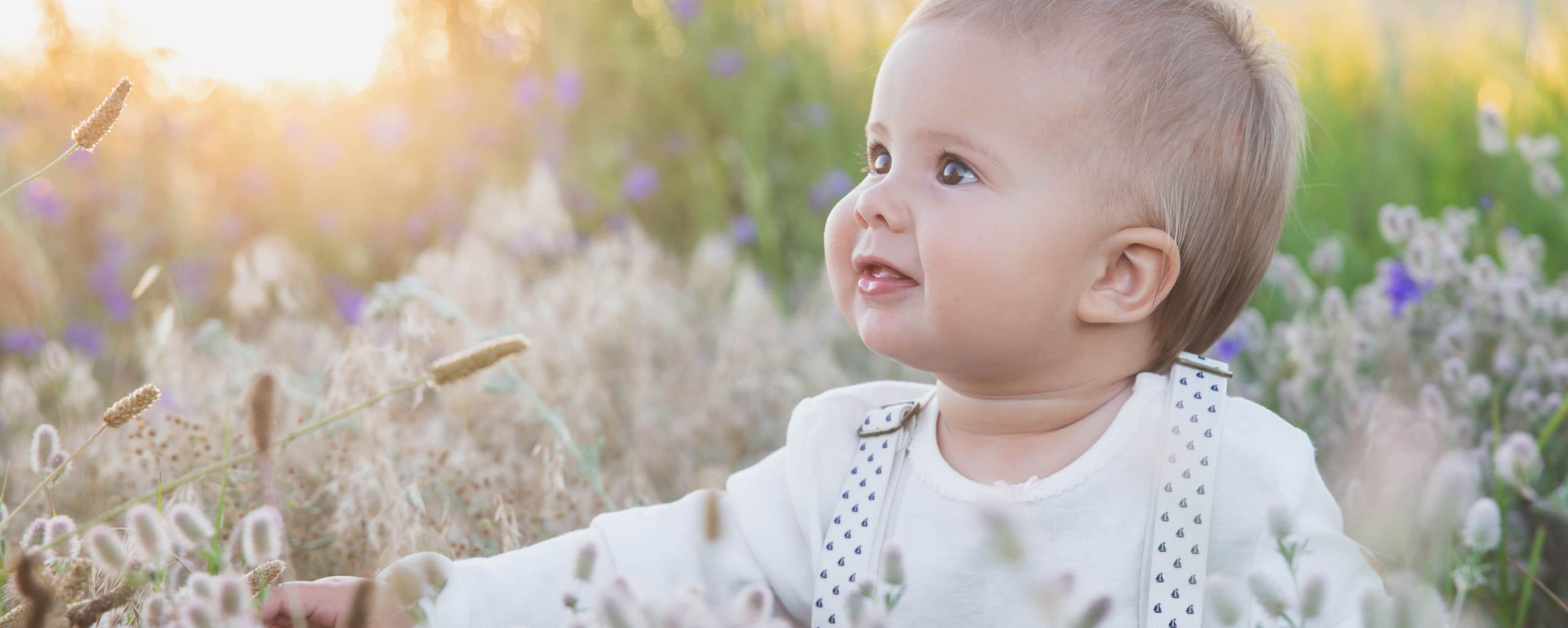 Baby mit hellem Haar sitzt in einer Wiese mit Gräsern und lila Blüten, trägt ein weißes Oberteil mit Hosenträgern und schaut lächelnd nach rechts.