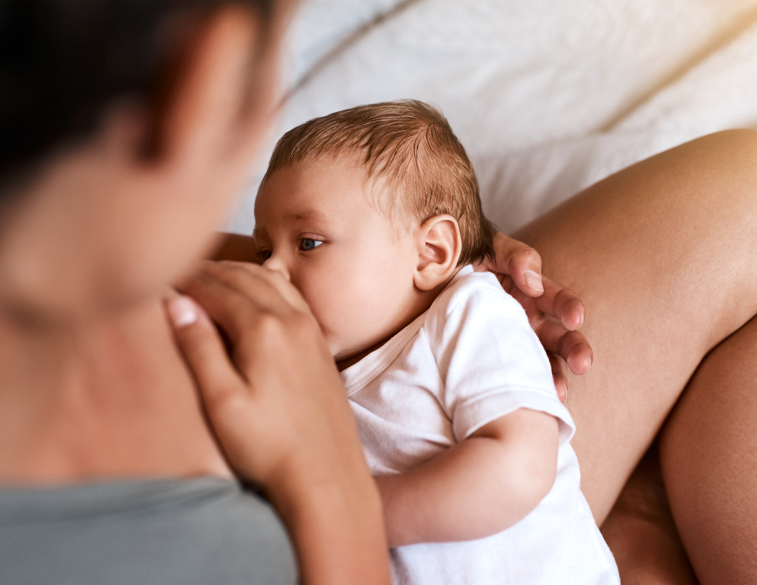 Nahaufnahme eines Babys in weißem Body, das an der Brust einer Person gestillt wird.