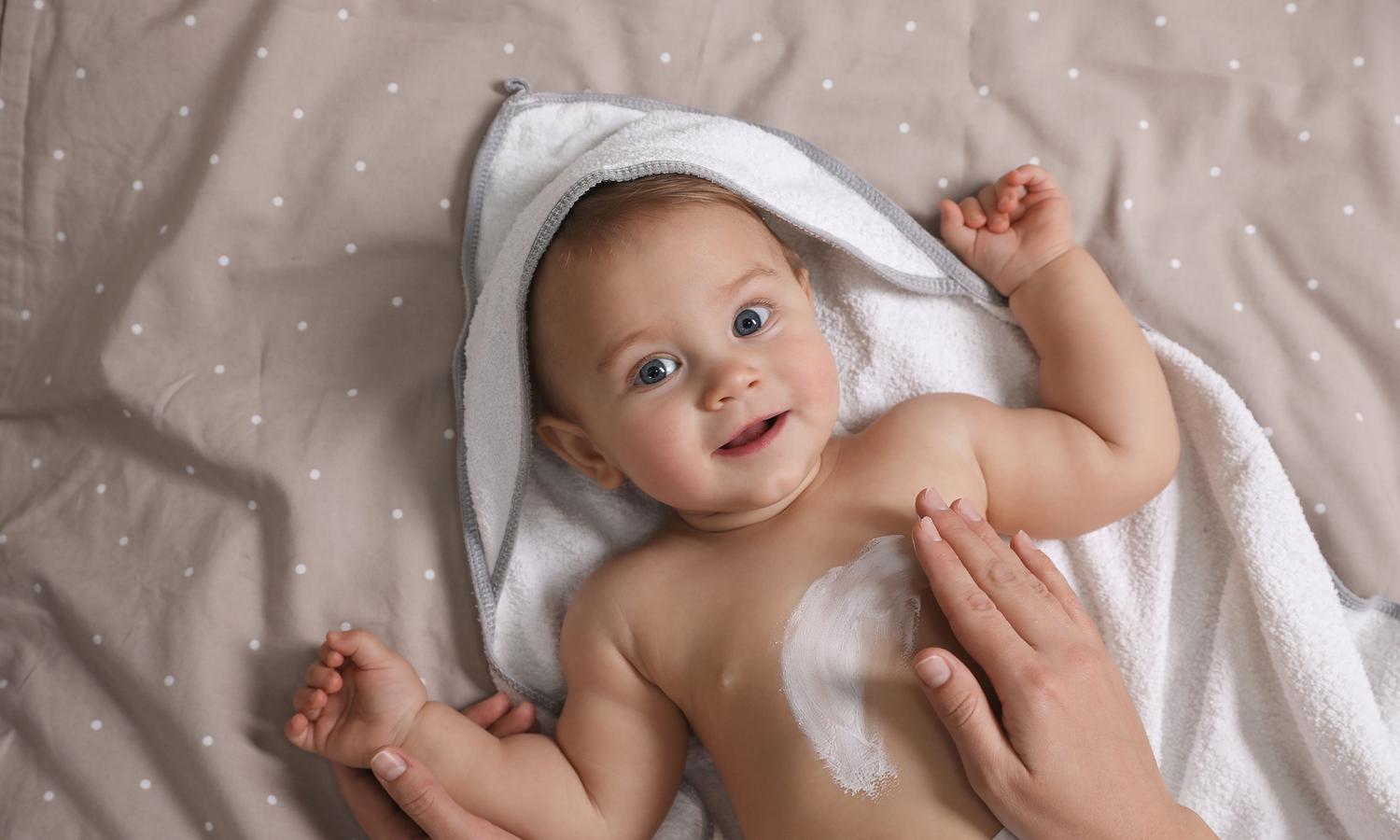 Baby liegt im Bademantel auf dem Rücken während eine Hand den Körper eincremt.